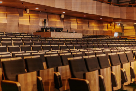 Interior view of a modern auditorium with empty rows of wooden seats, black cushions, and wooden...