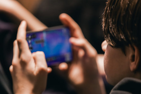 Young boy engages with a smartphone displaying vibrant game graphics, seated in a softly lit indoor...