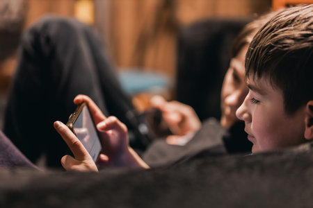 Two kids relax on a couch, one focused on a smartphone screen while the other interacts with a...