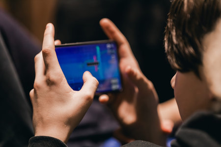 Young boy engages with mobile video game on smartphone, holding it with both hands, seated in a...