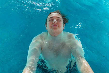 Young man swims underwater with arms outstretched in clear blue water, sunlight creates reflections and highlights on the surface aboveの写真素材