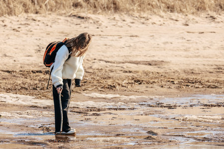 Child wearing a backpack inspects the wet sand on a beach at low tide, with grass and dunes in the...