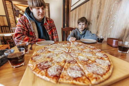 Adult man and young boy sit at a wooden table with a large pepperoni pizza, glasses of dark beverage, and empty plates in a restaurant interior