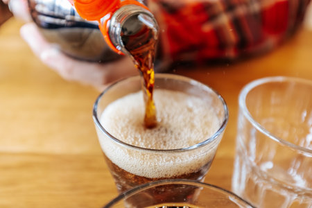 Soda is being poured into a clear glass with ice, while other empty glasses are placed on a wooden table in a casual dining environment