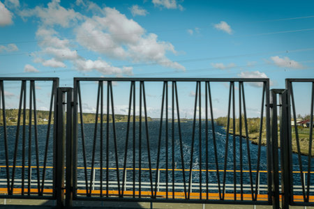 Metal railing features a geometric design, providing a view of the river and surrounding landscape under a clear blue sky with scattered clouds