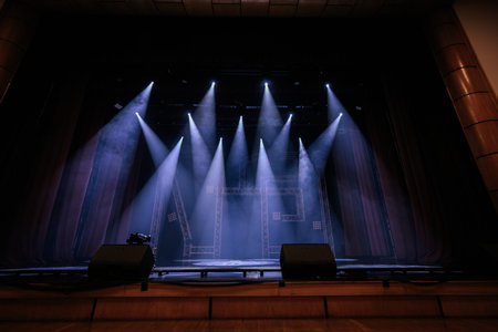 Stage area with bright spotlights casting beams in a dark theater, showcasing wooden flooring and draped curtains, prepared for an upcoming performance