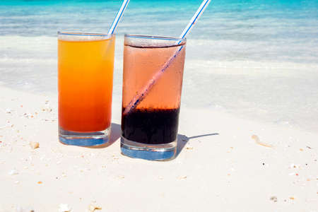 Two colorful cocktails laying on white sand, small corals covered beach in front of the amazing turquoise water of the indian ocean.の写真素材
