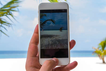 Hand of a caucasian male holding a smartphone and trying to capture the beautiful beach of the...