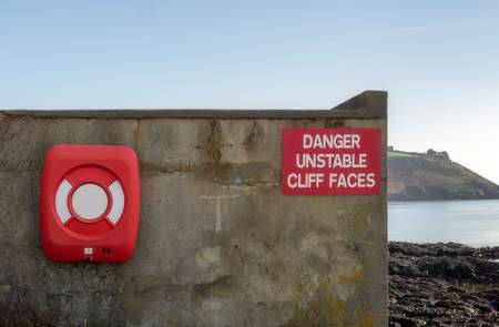 Fragment of a wall of the building standing on the side of the cliff on which hangs a red warning sign and a lifebuoy in the plastic box.の写真素材