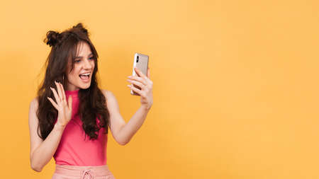 A smiling woman holds a smartphone on which she records a video message or talks in a video chat, standing on a yellow background. A copy of the space. Space for textの写真素材