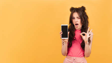 A smiling woman holds a smartphone with a clear screen, standing on a yellow background. A copy of the space. Space for textの写真素材