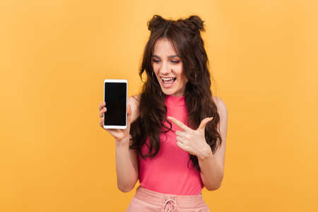 A smiling woman points to the blank screen of her smartphone, standing on a yellow background. A copy of the space. Space for textの写真素材