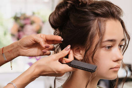 Shooting in a beauty salon. A hair stylist corrects the hairstyle of a young dark-haired girl with a hair comb.の写真素材