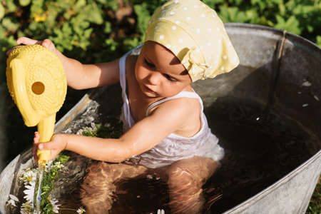 A little girl in a headscarf and with a watering can is bathing in the children's bathroom in the garden, on a hot sunny summer day.の写真素材