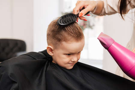 Shooting in a beauty salon. The hairdresser does the styling of the little boy with a hair dryer and a comb.の写真素材