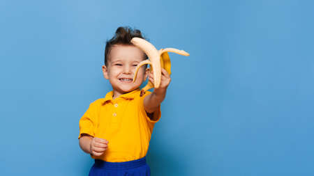 Happy portrait of a child in a yellow T-shirt holding a peeled banana for food, studio shot isolated on a blue backgroundの写真素材