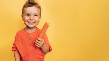 Studio shot of a smiling boy holding a fresh carrot on a yellow background. The concept of healthy baby food, carotene. A place for your text.の写真素材