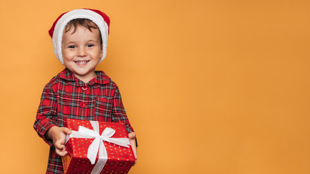 Studio photo of a baby boy in Christmas pajamas and a hat on a yellow background with a bright red gift box in his hands. A place for your text, advertising.の写真素材