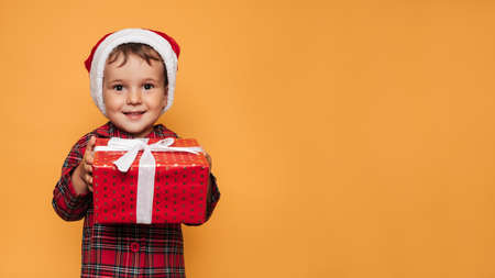 Studio photo of a baby boy in Christmas pajamas and a hat on a yellow background with a bright red gift box in his hands. A place for your text, advertising.の写真素材