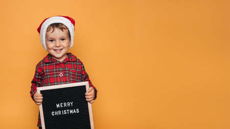 A smiling boy in a red Santa hat and pajamas, with a board with white letters and the inscription Merry Christmas in his hands. A place for your text and advertising.の写真素材