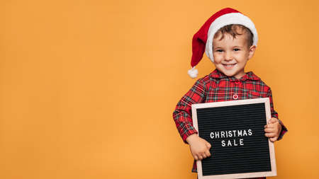 Studio photo of a baby boy in Christmas pajamas and a hat on a yellow background with a letter board with the text Merry Christmas in his hands. A place for your text, advertising.の写真素材