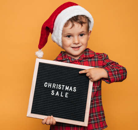Studio photo of a baby boy in Christmas pajamas and a hat on a yellow background with a letter board with the text Merry Christmas in his hands.の写真素材