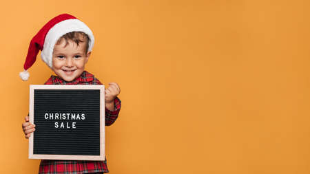 A smiling boy in a red Santa hat and pajamas, with a board with white letters and the inscription Merry Christmas in his hands. A place for your text and advertising.の写真素材