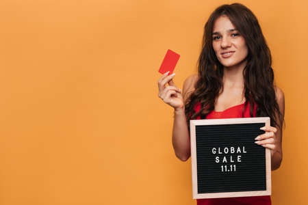 A smiling Caucasian woman holding a credit card and a message board with the inscription on it Global sale on November 11, isolated on a yellow background with a place to copy.の写真素材