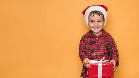 Studio photo of a baby boy in Christmas pajamas and a hat on a yellow background with a bright red...
