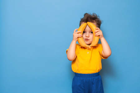 A boy in a yellow T-shirt is holding a banana with a smile. Funny expressions. The concept of nutrition. Fresh banana. Yellow style. Blue backgroundの写真素材