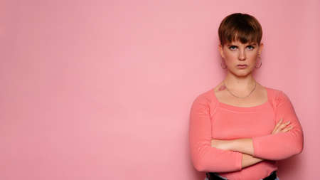 An irritated young woman with her arms crossed over her chest and looking at the camera with a skeptical look is isolated on a pink background. Human facial expressions. Body language. Copying Spaceの写真素材