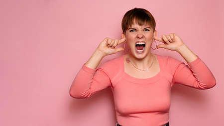 An angry, irritated young woman, isolated on a pink background, covers her ears from loud noises and screams.Human facial expressions, emotions. Body language. Copying space.の写真素材
