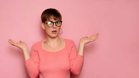 A young woman isolated on a pink background expresses bewilderment by spreading her hands. Human facial expressions, emotions. Body language. Copying space.の写真素材
