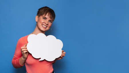 Studio shot of a smiling young woman holding a white empty banner in the form of a cloud on a blue background. Copy space, space for your ad or text.の写真素材
