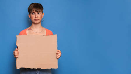 Studio shot of a young woman with a skeptical expression holding a blank cardboard sheet on a blue background. Copy space, space for your ad or text.の写真素材