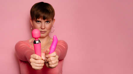 Studio shot of a young woman holding two pink female vibrators. Bright pink background. A place for your text or advertisement. The concept of female orgasmの写真素材