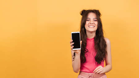 A smiling woman holds a smartphone with a clear screen, standing on a yellow background.の写真素材