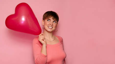 Portrait of a bright young woman with a red heart-shaped balloon on a pink background. A place for your text.の写真素材