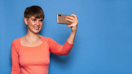 Portrait of a young woman taking a selfie on a smartphone on a blue background. A place for your text. The concept of video communication at a distanceの写真素材