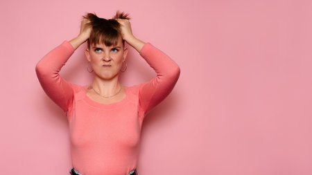 An angry, irritated young woman, isolated on a pink background, holds her head with her hands, feeling a headache.Human facial expressions, emotions. Body language. Copying space.の写真素材