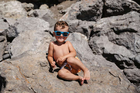 A child in sunglasses is sitting and sunbathing on large boulders, on the beach near the sea.の写真素材