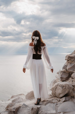 A young woman in a white dress stands barefoot on a cliff face in full height. The dress flutters in the wind. A sacred glow breaks through the clouds. The girl looks like an angelの写真素材