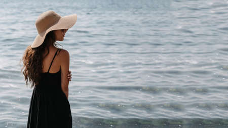 A picture of a beautiful young woman with long dark hair in a dress and a straw hat, posing on the beach, standing with her back and looking at the endless sea. A place for your text.の写真素材