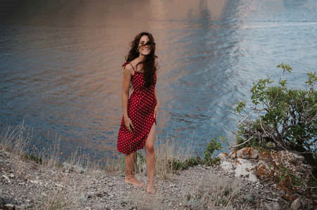 A picture of a young Caucasian woman in a red dress at the edge of a cliff near the beach and the sea with a sincere smile on her face and tangled hair from the wind. The concept of freedom and travelの写真素材