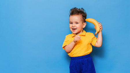 Funny face with a banana. A boy in a yellow T-shirt is holding a banana with a smile. Funny expressions. The concept of nutrition. Fresh banana. Yellow style. Blue backgroundの写真素材