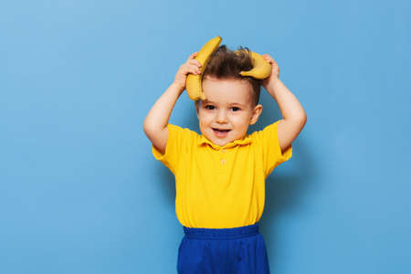 A boy in a yellow T-shirt is holding a banana with a smile. Funny expressions. The concept of nutrition. Fresh banana. Yellow style. Blue backgroundの写真素材