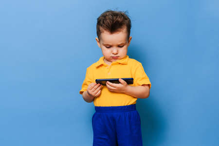 A little boy looks at the screen of a mobile phone. Studio photo on a blue background.の写真素材