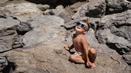 A child in sunglasses is sitting and sunbathing on large boulders, on the beach near the sea.の写真素材