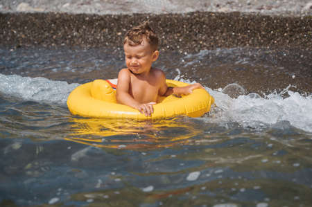 Baby boy splashes water in the sea, floating in a yellow inflatable circle. The kid is having fun and fooling aroundの写真素材