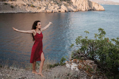 A young attractive Caucasian woman in a red dress at the edge of a cliff near the beach and the sea raised her hands up to the sun. The concept of freedom and travelの写真素材